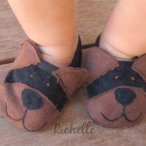 May include: Baby booties shaped like dog faces. The booties are brown and black, with stitched details for the eyes, nose, and mouth. The booties are made of soft material and are designed to fit a baby's feet.