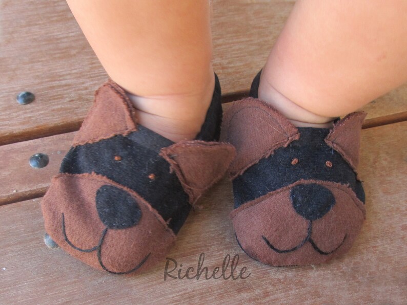 May include: Baby booties shaped like dog faces. The booties are brown and black, with stitched details for the eyes, nose, and mouth. The booties are made of soft material and are designed to fit a baby's feet.