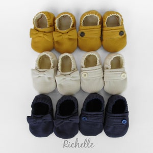 May include: Three pairs of baby shoes in different colours: mustard yellow, beige, and navy blue. Each pair has a bow on the front and a button closure on the side. The shoes are made of a soft, linen-like fabric.