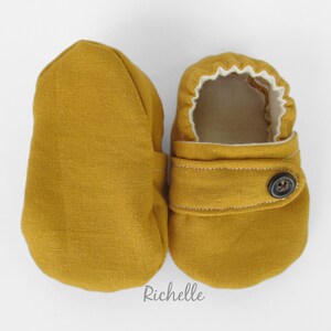 May include: A pair of mustard yellow baby booties with a button closure and white lining. Richelle is written on the bottom of the image.