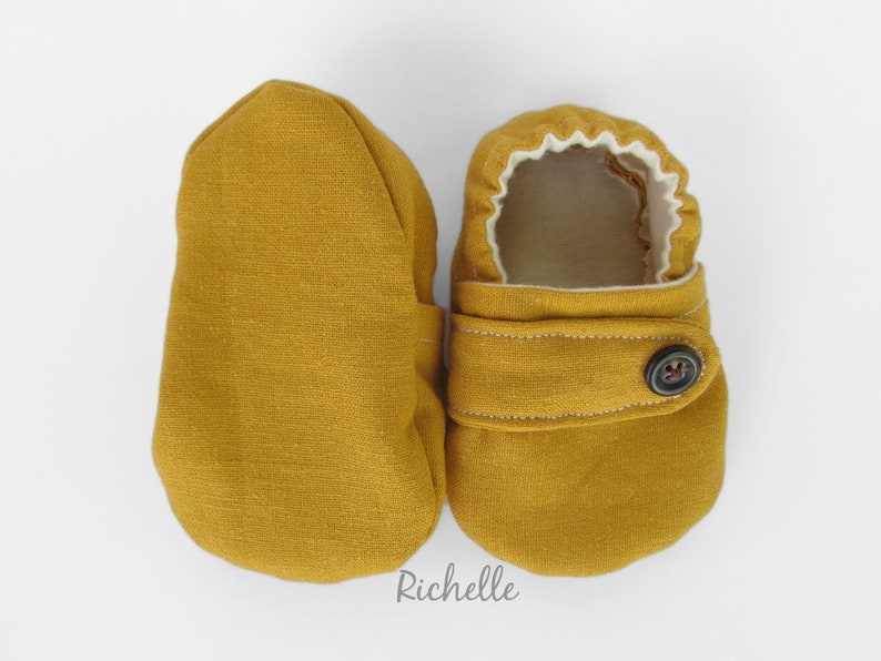 May include: A pair of mustard yellow baby booties with a button closure and white lining. Richelle is written on the bottom of the image.
