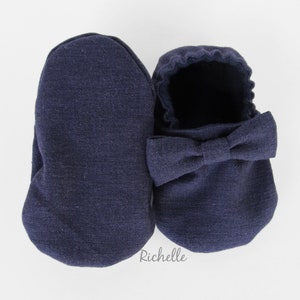 May include: A pair of navy blue baby booties with a bow on the top. The booties are made of a soft, textured fabric.