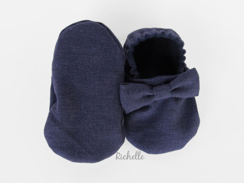 May include: A pair of navy blue baby booties with a bow on the top. The booties are made of a soft, textured fabric.