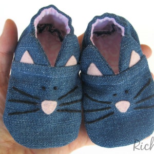 May include: A pair of blue denim baby shoes with pink felt cat ears and black felt whiskers.
