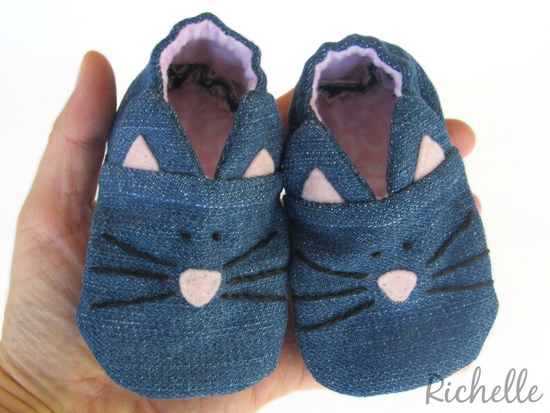 May include: A pair of blue denim baby shoes with pink felt cat ears and black felt whiskers.