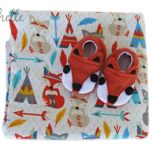 May include: A white baby blanket with a pattern of red, blue, and yellow foxes, teepees, and arrows. Two red fox baby booties are on top of the blanket.