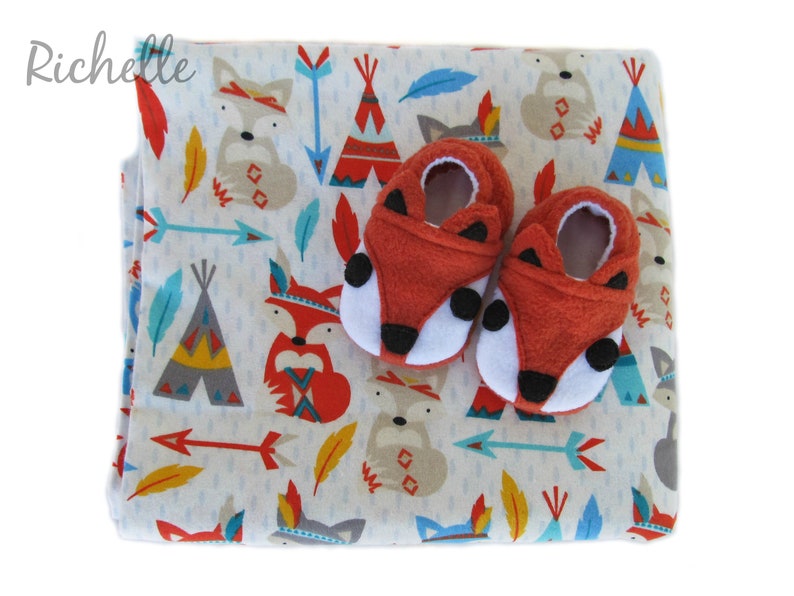 May include: A white baby blanket with a pattern of red, blue, and yellow foxes, teepees, and arrows. Two red fox baby booties are on top of the blanket.