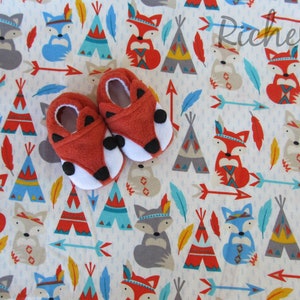 May include: A pair of red fox baby booties with white fur trim and black eyes. The booties are on a white background with a repeating pattern of blue, red, and yellow teepees, foxes, and arrows.