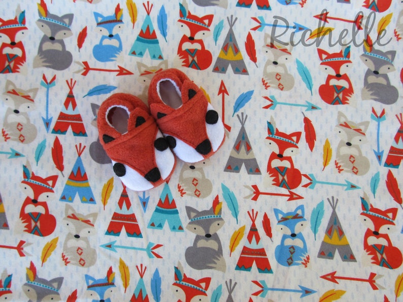 May include: A pair of red fox baby booties with white fur trim and black eyes. The booties are on a white background with a repeating pattern of blue, red, and yellow teepees, foxes, and arrows.