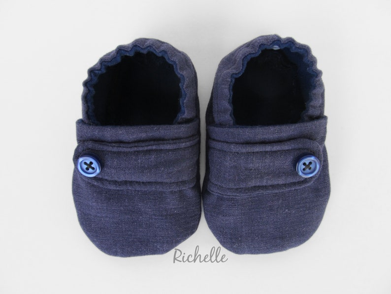 May include: A pair of dark blue baby booties with a button closure. The booties are made of a soft, textured fabric.