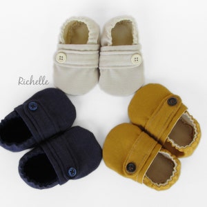 May include: Three pairs of baby shoes in different colors: beige, navy blue, and mustard yellow. Each pair has a single button closure.