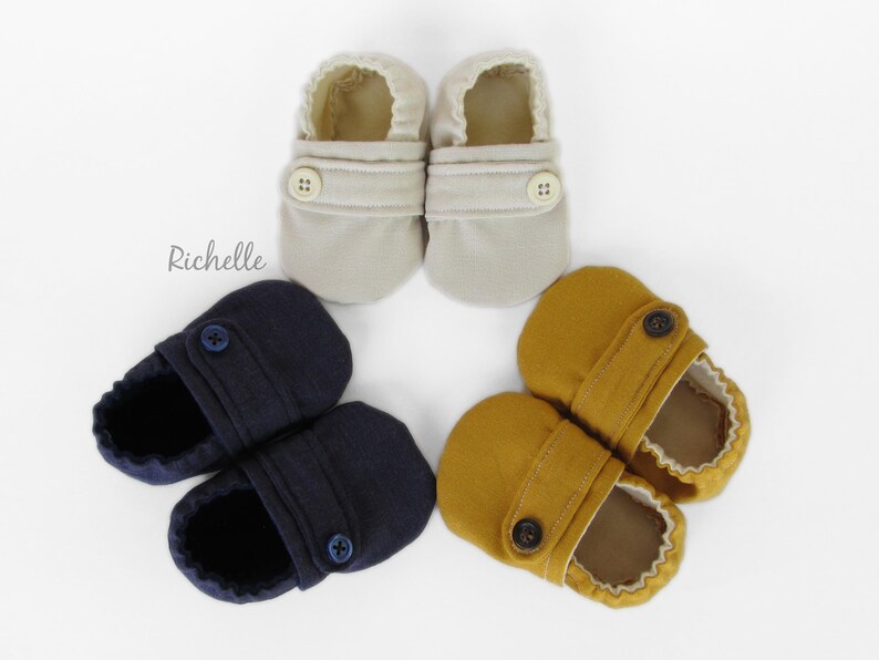 May include: Three pairs of baby shoes in different colors: beige, navy blue, and mustard yellow. Each pair has a single button closure.