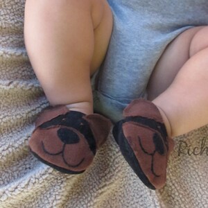 May include: A close-up of a baby's feet wearing brown and black dog-faced booties. The booties have a black nose and ears. The baby is wearing a gray onesie. The background is a light brown, textured surface.