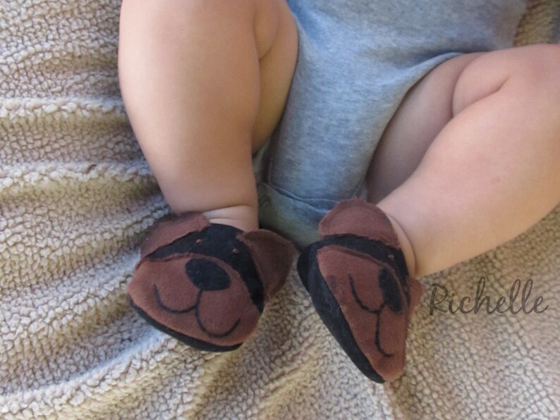 May include: A close-up of a baby's feet wearing brown and black dog-faced booties. The booties have a black nose and ears. The baby is wearing a gray onesie. The background is a light brown, textured surface.