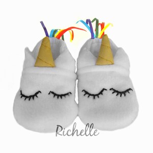 May include: White baby booties with a unicorn design. The booties have a gold horn, black eyelashes, and a rainbow ribbon.