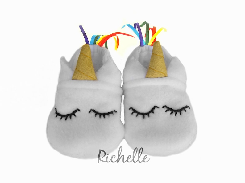 May include: White baby booties with a unicorn design. The booties have a gold horn, black eyelashes, and a rainbow ribbon.