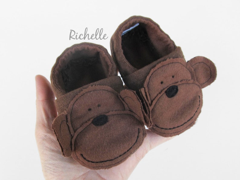 May include: A pair of brown baby booties with monkey faces. The booties have brown felt ears and black eyes and noses.