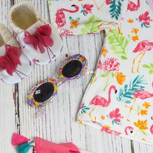 May include: A set of baby shoes with pink bows and a flamingo print, sunglasses with a colourful frame, and a white t-shirt with a flamingo and floral print.