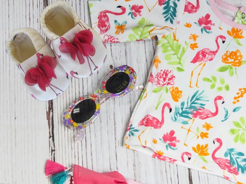 May include: A set of baby shoes with pink bows and a flamingo print, sunglasses with a colourful frame, and a white t-shirt with a flamingo and floral print.