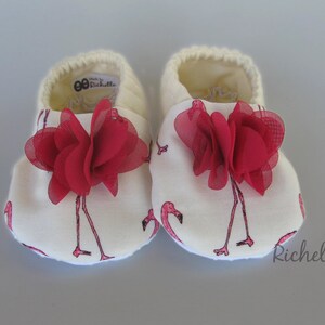 May include: A pair of white baby shoes with a pink flamingo print and red fabric flower accents. The shoes have the text "Made by Richelle" on the back.