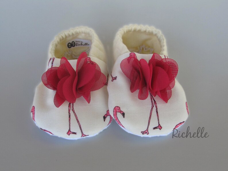 May include: A pair of white baby shoes with a pink flamingo print and red fabric flower accents. The shoes have the text "Made by Richelle" on the back.