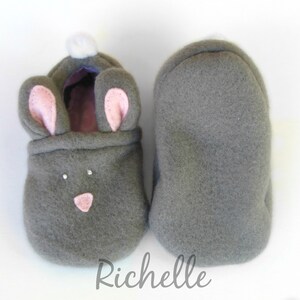 May include: A pair of gray fleece baby booties shaped like bunnies. One bootie features pink inner ears, a pink nose, and two small rhinestones. The other bootie is plain gray. A white pom-pom is on the back of the bunny bootie.