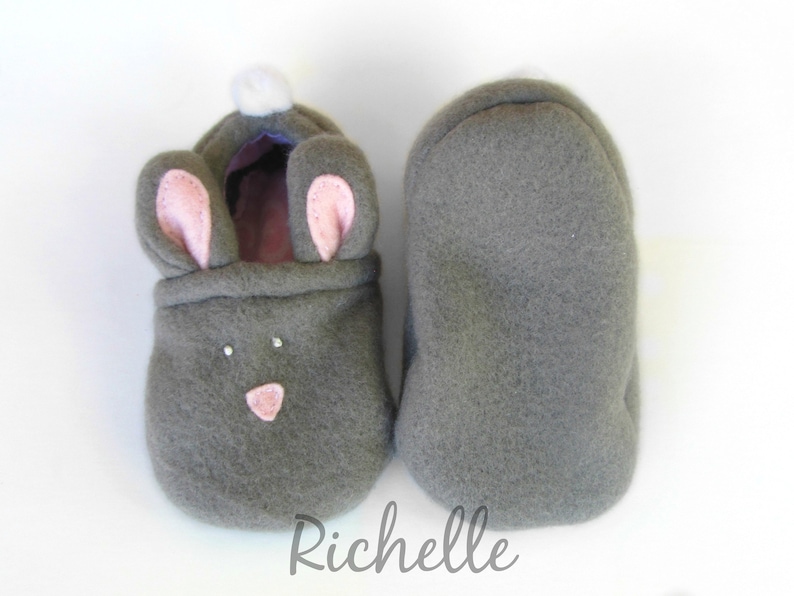 May include: A pair of gray fleece baby booties shaped like bunnies. One bootie features pink inner ears, a pink nose, and two small rhinestones. The other bootie is plain gray. A white pom-pom is on the back of the bunny bootie.