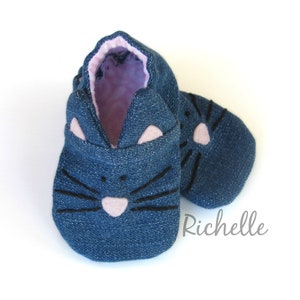 May include: A pair of blue denim baby booties with a cat face design. The cat face features black whiskers, a pink nose, and pink ears.