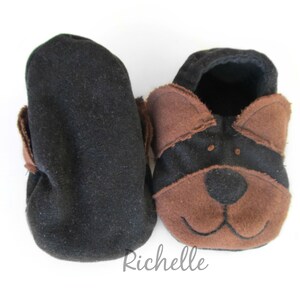 May include: A pair of baby booties, one black and one with a dog face design. The dog face features brown ears, a brown snout, and a black face. The booties are made of soft fabric and are designed for infants.