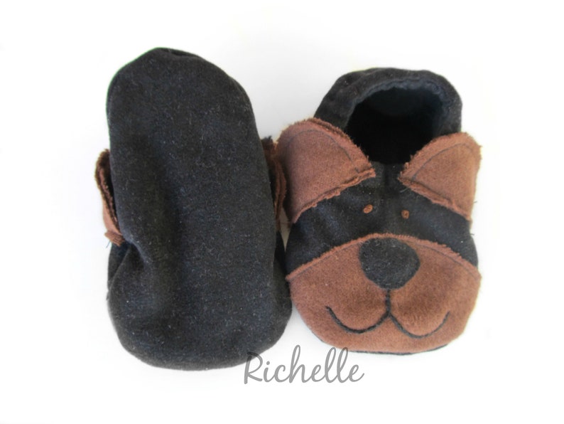 May include: A pair of baby booties, one black and one with a dog face design. The dog face features brown ears, a brown snout, and a black face. The booties are made of soft fabric and are designed for infants.