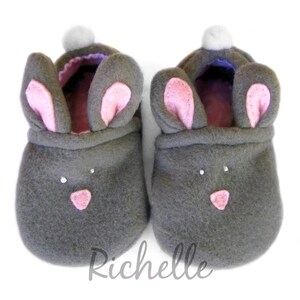 May include: A pair of gray bunny slippers with pink inner ears and noses. Each slipper has a white pom-pom on top and small white accents for eyes. The slippers are designed to resemble a bunny.