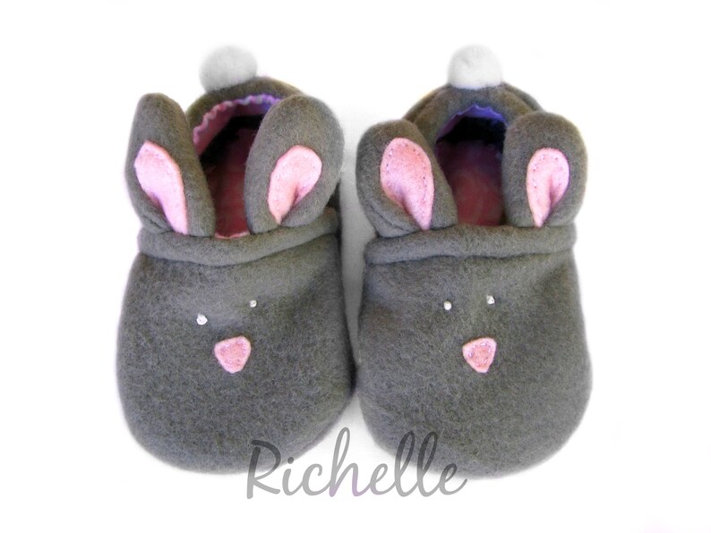 May include: A pair of gray bunny slippers with pink inner ears and noses. Each slipper has a white pom-pom on top and small white accents for eyes. The slippers are designed to resemble a bunny.