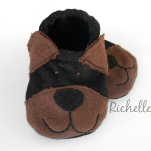 May include: A pair of baby booties in the shape of a dog's face. The booties are primarily brown and black, with the dog's features, including ears, eyes, nose, and mouth, carefully stitched onto the fabric. The booties are designed for infants.
