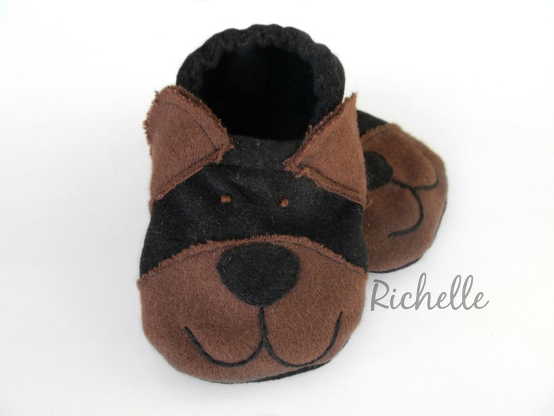 May include: A pair of baby booties in the shape of a dog's face. The booties are primarily brown and black, with the dog's features, including ears, eyes, nose, and mouth, carefully stitched onto the fabric. The booties are designed for infants.