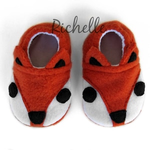 May include: A pair of orange fleece baby booties with a fox face design. The booties have black eyes, a black nose, and white fur around the eyes.