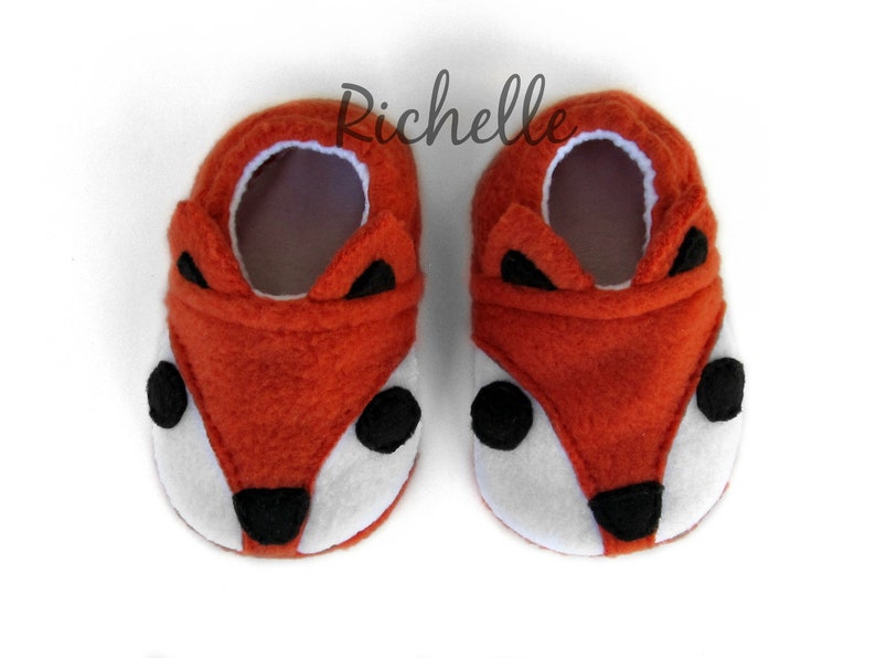 May include: A pair of orange fleece baby booties with a fox face design. The booties have black eyes, a black nose, and white fur around the eyes.