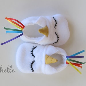 May include: White baby booties with a unicorn design. The booties have a gold horn, black embroidered eyes, and a rainbow ribbon tail.