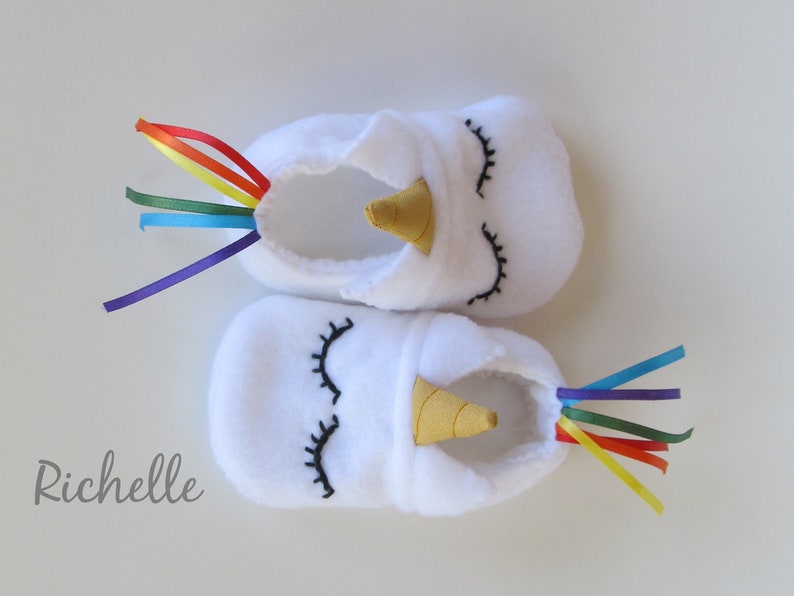 May include: White baby booties with a unicorn design. The booties have a gold horn, black embroidered eyes, and a rainbow ribbon tail.