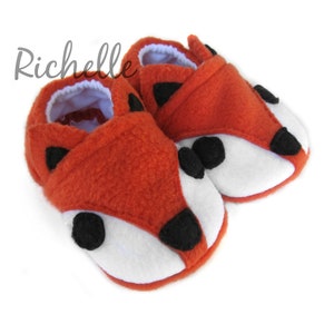 May include: A pair of orange fleece baby booties with white and black fox faces.