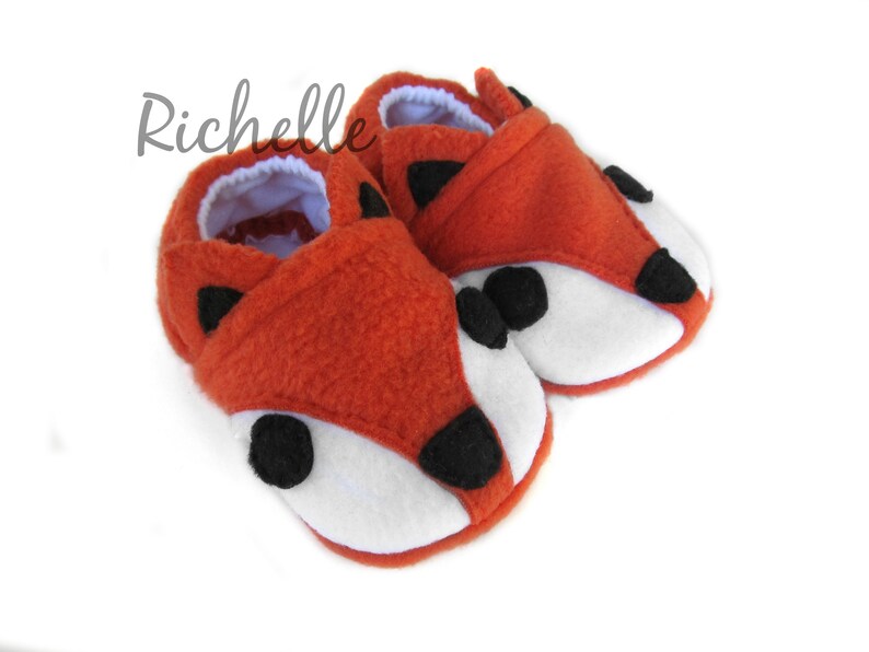 May include: A pair of orange fleece baby booties with white and black fox faces.