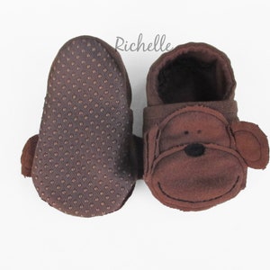 May include: A pair of brown baby booties with a monkey face design. The booties have a brown fleece exterior and a brown textured sole.