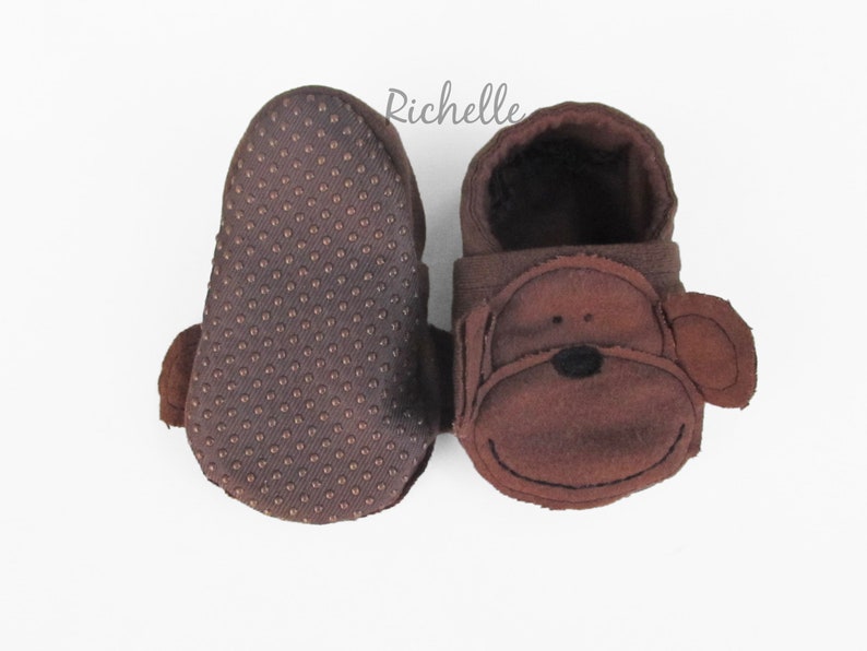 May include: A pair of brown baby booties with a monkey face design. The booties have a brown fleece exterior and a brown textured sole.