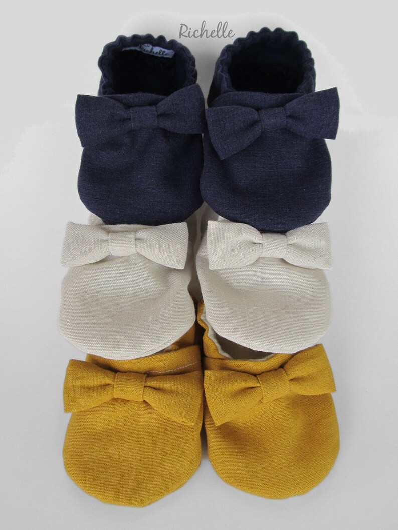 May include: Three pairs of baby shoes with bows. The top pair is navy blue, the middle pair is cream, and the bottom pair is mustard yellow.