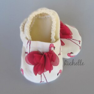 May include: A pair of white baby booties with a pink flamingo print and a red bow. The booties are lined with a soft, white material.