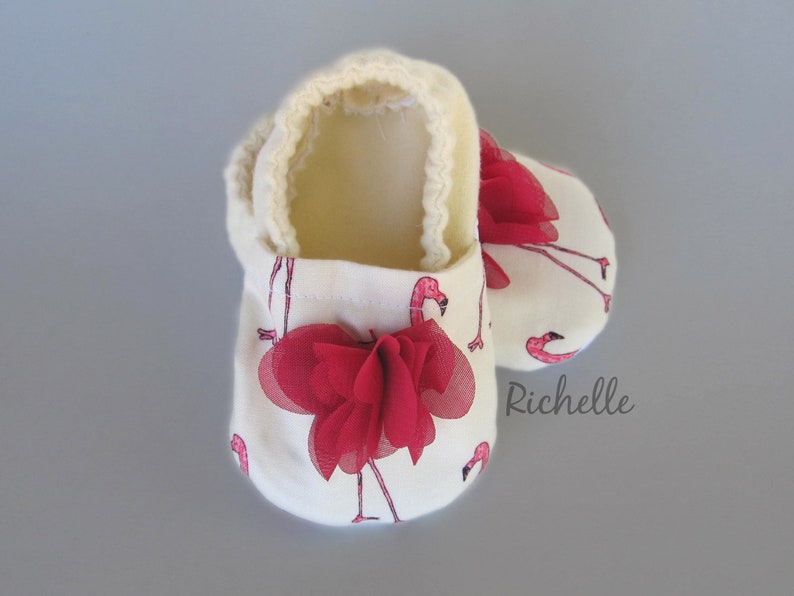 May include: A pair of white baby booties with a pink flamingo print and a red bow. The booties are lined with a soft, white material.
