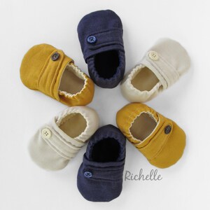 May include: Six pairs of baby shoes in three colors: navy blue, beige, and mustard yellow. Each shoe has a single button closure and a soft, textured fabric.