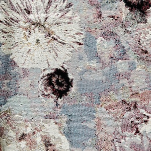 May include: A close-up of a tapestry featuring a floral design. The tapestry is woven from a textured fabric. The colours in the tapestry are muted and include shades of blue, brown, and white.