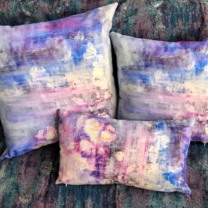 May include: Three decorative throw pillows with abstract watercolour designs in shades of purple, blue and white.