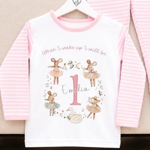 May include: A white long-sleeve shirt and matching pants set with pink and white horizontal stripes on the sleeves and legs. The shirt features a graphic of four mice in tutus, a swan, and the text "When I wake up I will be 1 Emilia".