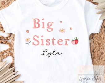 May include: White t-shirt with pink text that says "Big Sister Lyla" with pink strawberry and flower illustrations.